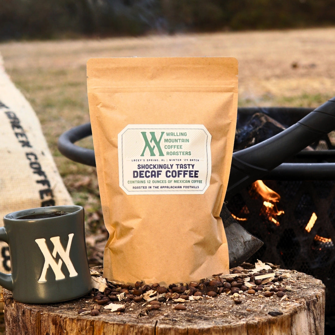 A 12oz bag of "Shockingly Tasty Decaf Coffee" from Walling Mtn. Coffee rests near a mug and fire pit, surrounded by coffee beans. Naturally decaffeinated, it's crafted using traditional roasting methods.