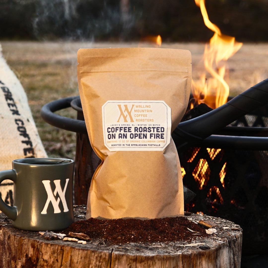 A 12oz Bag of Campfire Roast Coffee by Walling Mtn. Coffee and a mug sit on a tree stump by the campfire, with a sack of organic Colombian/Peruvian blend in the background, highlighting traditional roasting methods.