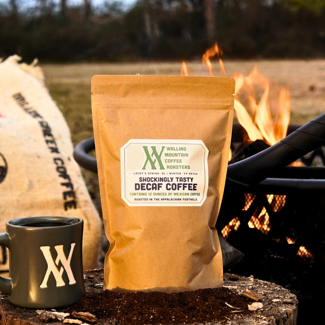 A 12oz Bag of Walling Mtn. Coffee's decaf coffee sits on soil next to a matching mug by the fire pit, with a bean sack in the background showcasing its traditional roasting methods.