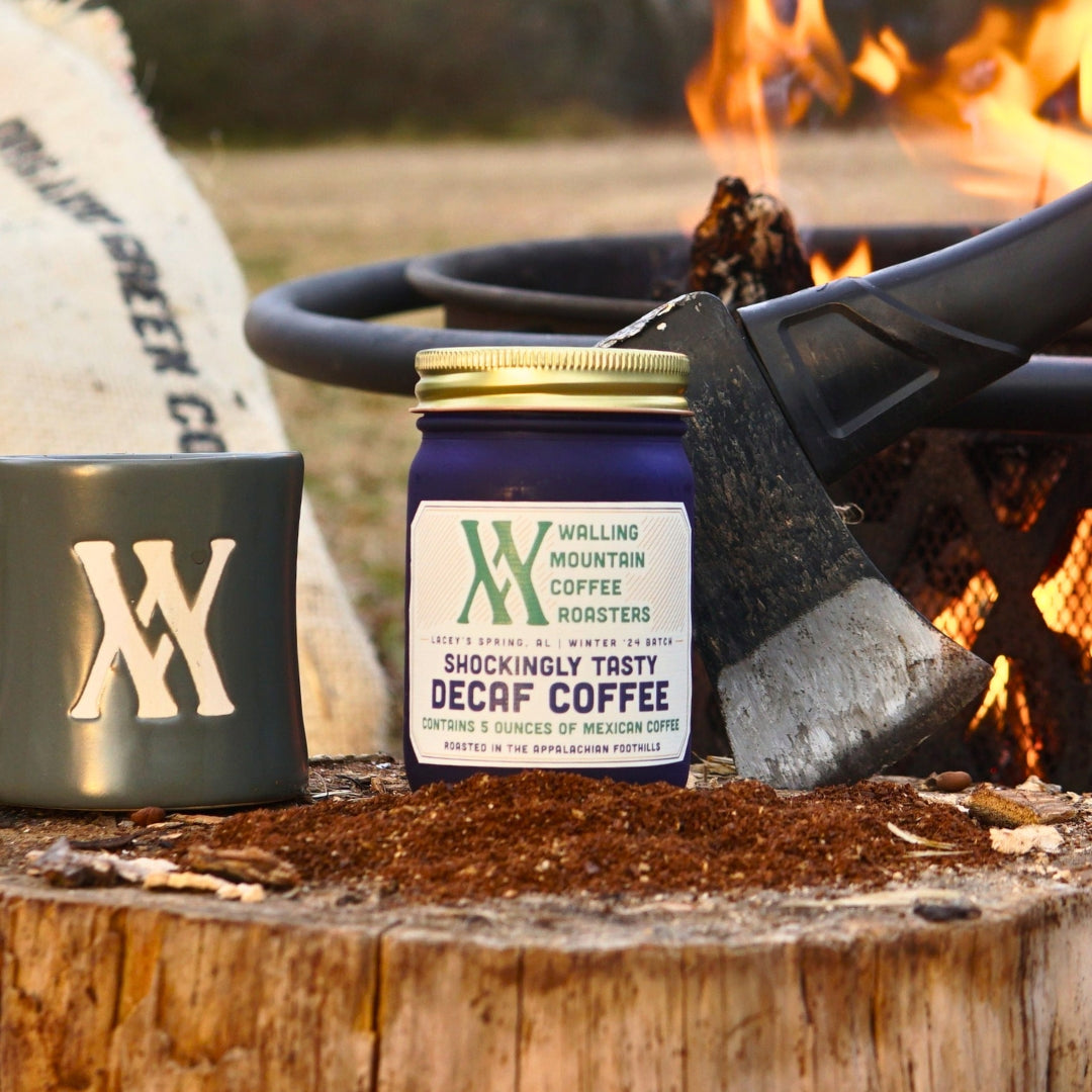 A 5oz jar of Decaf Coffee – Walling Mtn. and a mug rest on a stump near an axe and firepit, presenting the rich flavors of naturally decaffeinated Colombian coffee in a rustic setting.