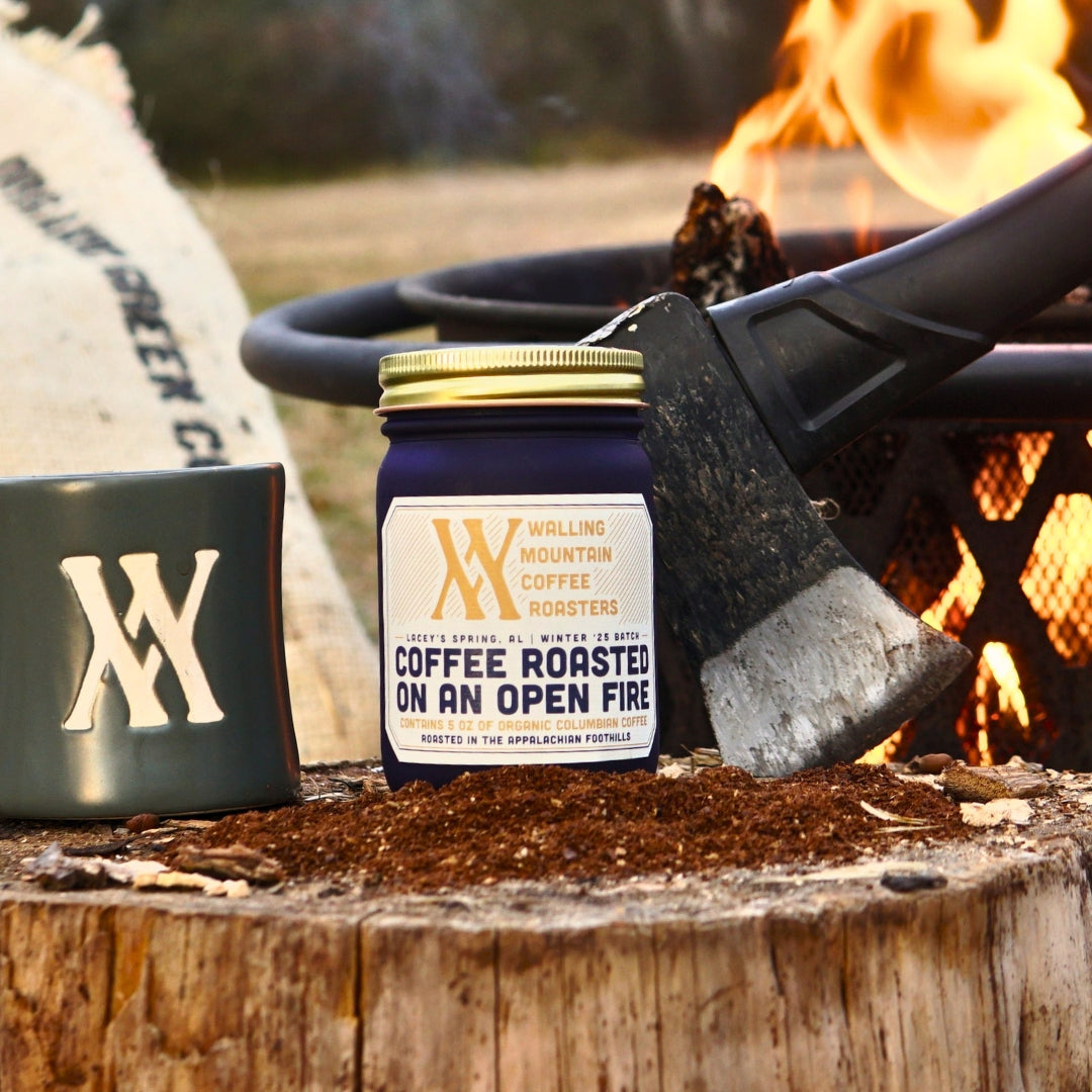 A 5oz Jar of Campfire Roast Coffee from Walling Mtn. sits on a tree stump beside a mug and an axe, with a smoldering fire pit in the background. This organic blend captures the rugged outdoor spirit, ideal for early mornings in nature.