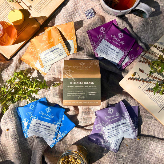 Packets of Piper & Leaf Tea Co.'s Wellness Blends, which include invigorating ginger and soothing herbal varieties, are elegantly arranged with a mug of tea on a textured cloth. Scattered herbs and dried flowers add charm, while an open book and honey jar complete the serene setting.