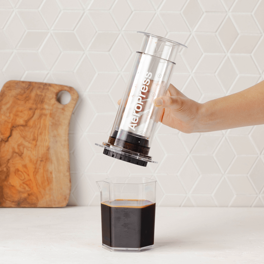 A hand presses an AeroPress Clear XL Portable Coffee Press by AeroPress over a glass of black coffee, with a wooden cutting board in the background.