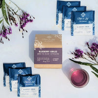 Piper & Leaf Blueberry Jubilee tea packages with a box and a cup of tea on a white background with purple flowers.