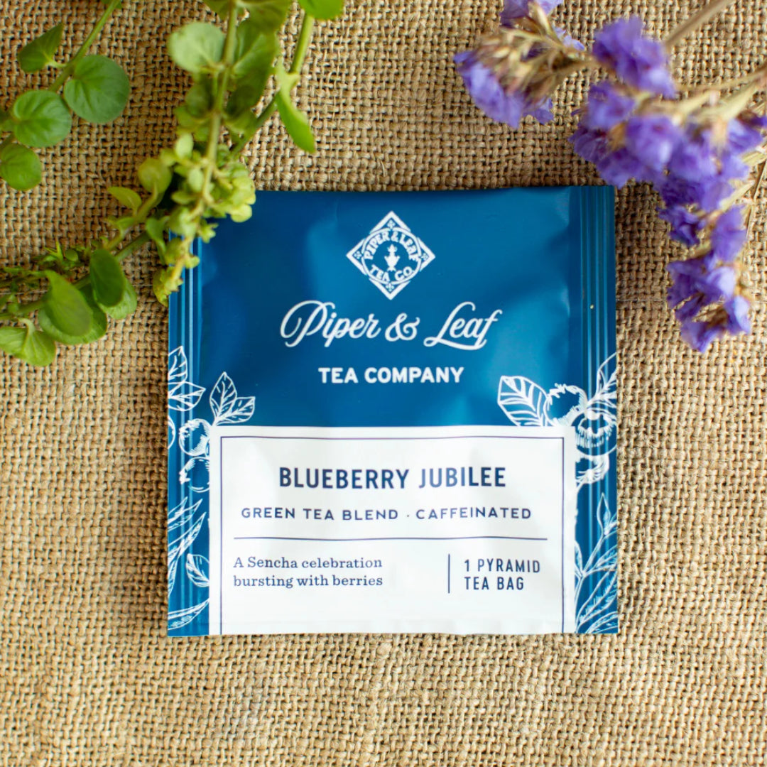 A Piper & Leaf Tea Co. Blueberry Jubilee box of 12 individually wrapped tea bag sachets rests on burlap amid green and purple foliage, showcasing its unique blueberry tea blend made with Sencha green tea.