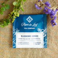 A Piper & Leaf Tea Co. Blueberry Jubilee box of 12 individually wrapped tea bag sachets rests on burlap amid green and purple foliage, showcasing its unique blueberry tea blend made with Sencha green tea.