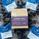 A box of Piper & Leaf Tea Co.'s Blueberry Jubilee—12 individually wrapped sachets—is displayed on a weathered wooden surface, surrounded by loose blueberries and five individual tea bags.