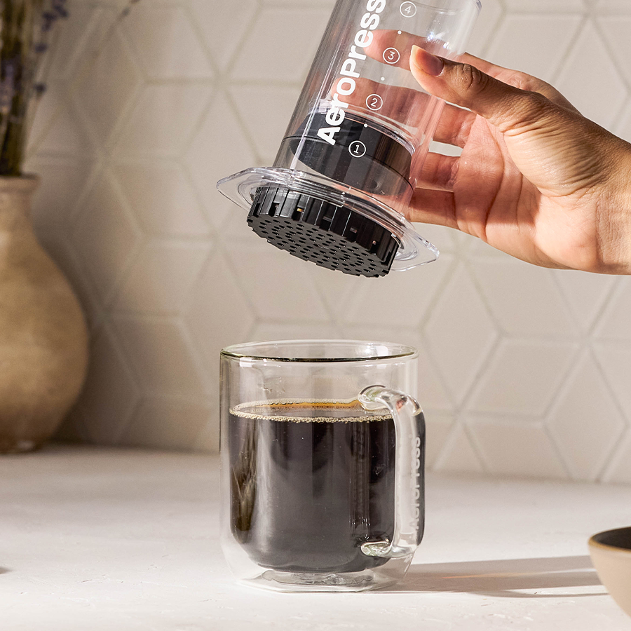 A hand lifts the AeroPress Clear Coffee Press 2 Min Portable Brew by AeroPress above a clear glass mug of coffee on a kitchen counter, showcasing its versatile 3-in-1 brew technology.