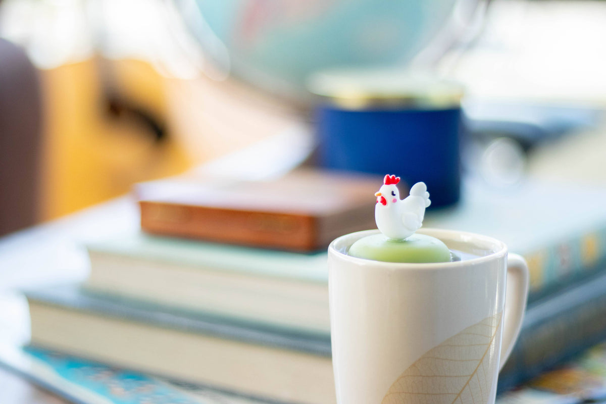 A white mug with a leaf design holds a drink topped with the Float2infuse Tea Infuser: Chicken - Floating Tea Infuser, perched charmingly on its lid. This delightful accessory rests atop stacked books, with a blurred background completing the cozy scene.