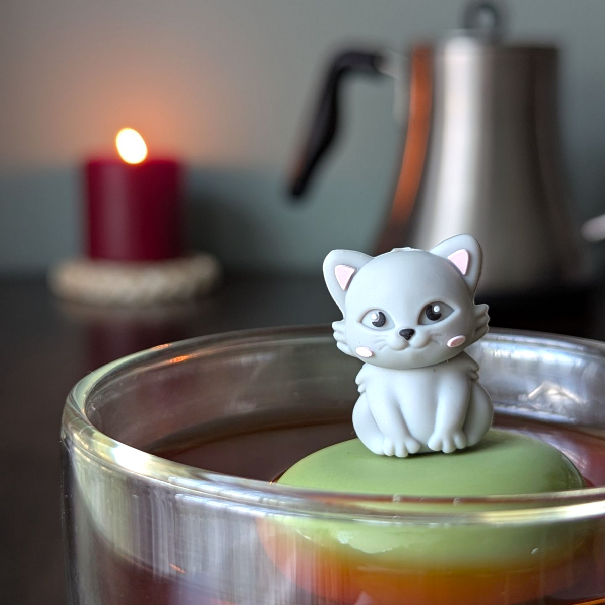 The Float2infuse Tea Infuser’s Grey Cat - Floating Tea Infuser rests on a green surface on top of a tea glass, with a glowing candle and metal kettle softly blurred in the background.