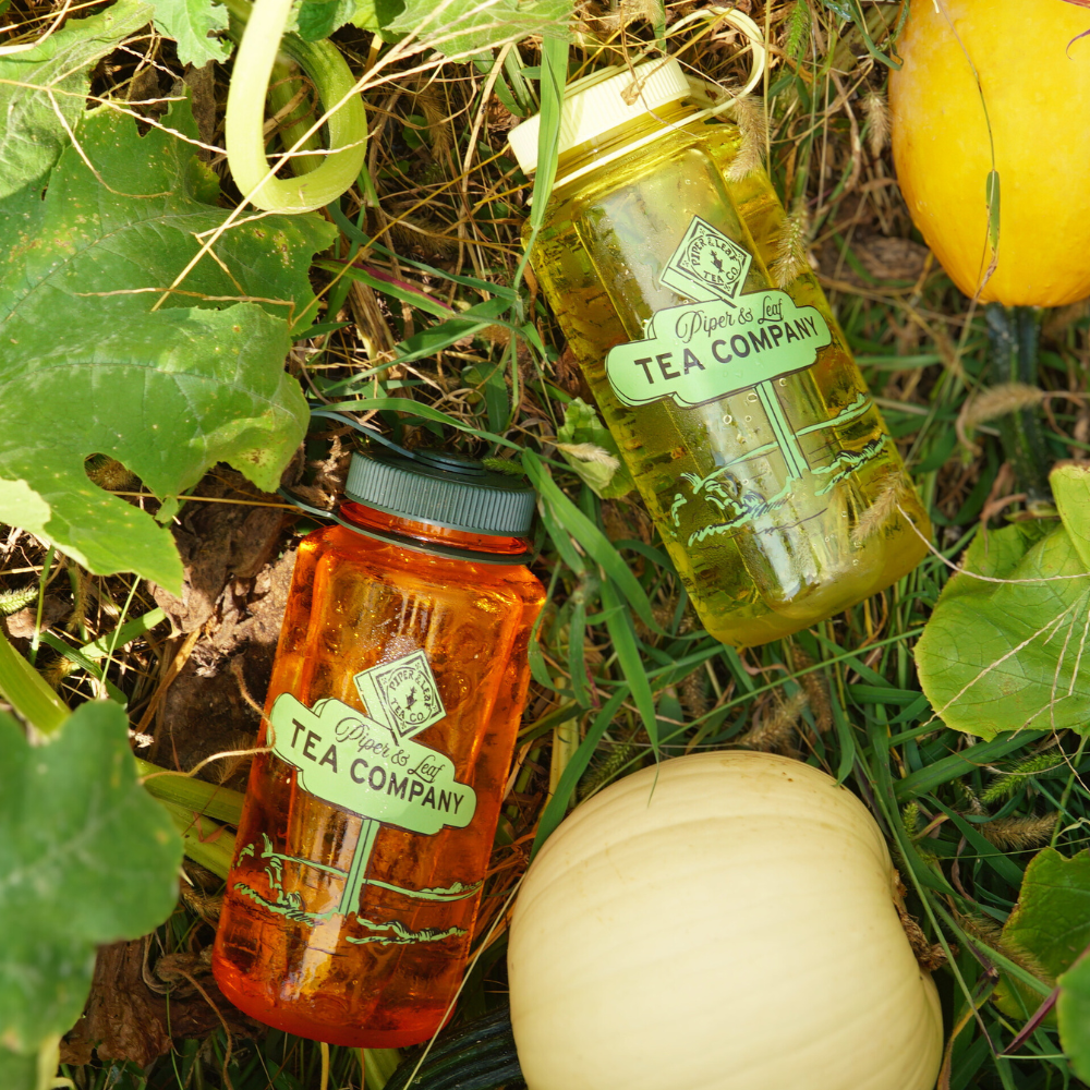 Two BPA-free Piper & Leaf Nalgene Water (Tea) Bottles with "Piper & Leaf Tea Co." labels, one filled with a green liquid and the other with an amber liquid, are placed on the ground among green leaves and squash.