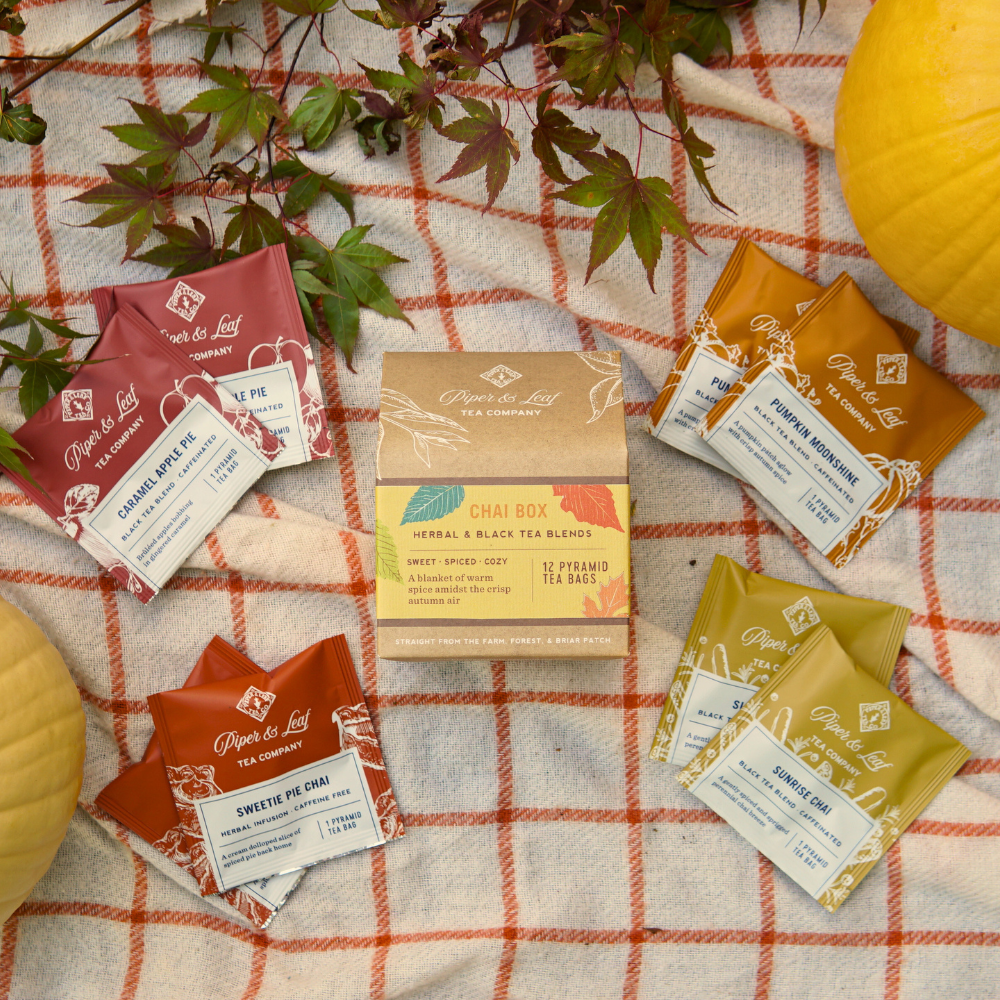 Piper & Leaf Tea Co.'s Chai Box, which includes 12 tea bags, is nestled among autumn-themed tea packets on a plaid cloth, accented by decorative leaves and pumpkins, exuding the rich aroma of autumn spices.