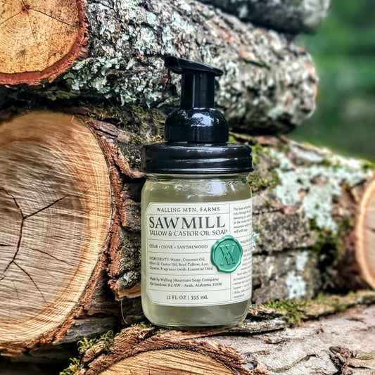 A bottle of Walling Mountain Farms Sawmill 12oz Tallow & Castor Oil Foaming Hand Soap sits on stacked logs outdoors, framed by trees and wood in the background.