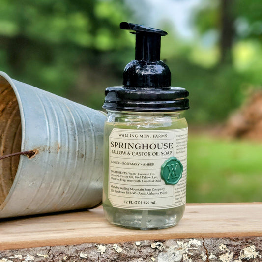 A bottle of Walling Mountain Farms Springhouse 12oz Tallow & Castor Oil Foaming Hand Soap sits on a wooden surface next to a metal bucket, outdoors with greenery in the background.