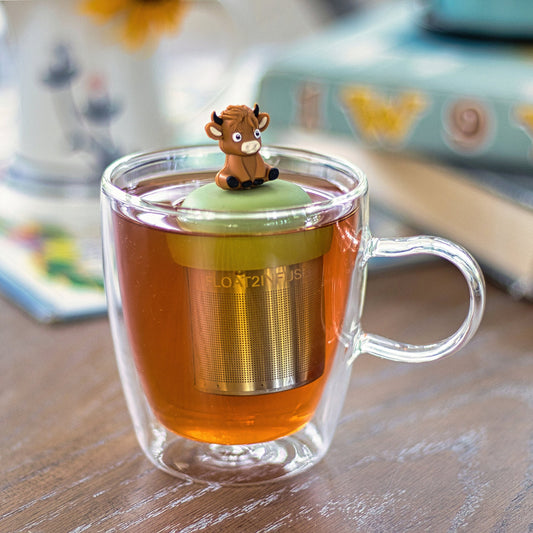 A glass cup of tea features the Float2infuse Tea Infuser’s “Baby Highland Cow - Floating Tea Infuser” nestled inside, its charming cartoon cow figurine perched on top. In the softly blurred background, books and a floral ornament add a cozy touch.