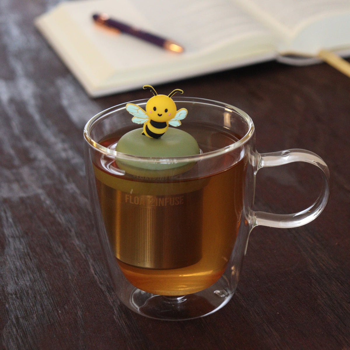 A glass mug of tea features the Float2infuse Tea Infuser’s Bumble Bee - Floating Tea Infuser, adorned with a whimsical bee-shaped topper; in the background, an open notebook and pen rest on a dark wooden table—making this reusable tea steeper a delightful and practical loose-leaf accessory for any cozy workspace.
