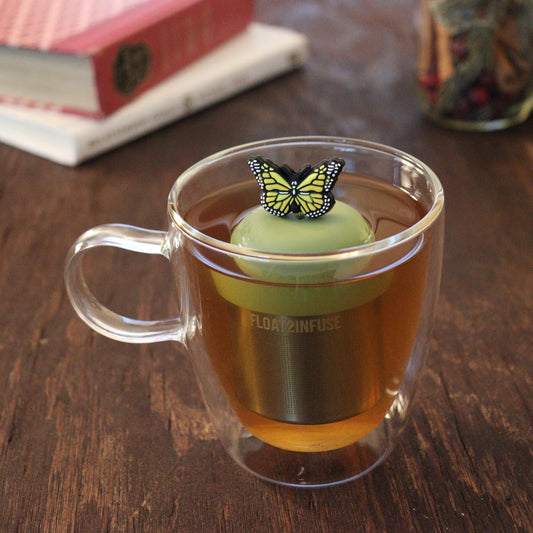 A glass mug of tea with the Float2infuse Tea Infuser, featuring the Yellow Butterfly design, sits on a wooden table while a book and a jar rest in the background.
