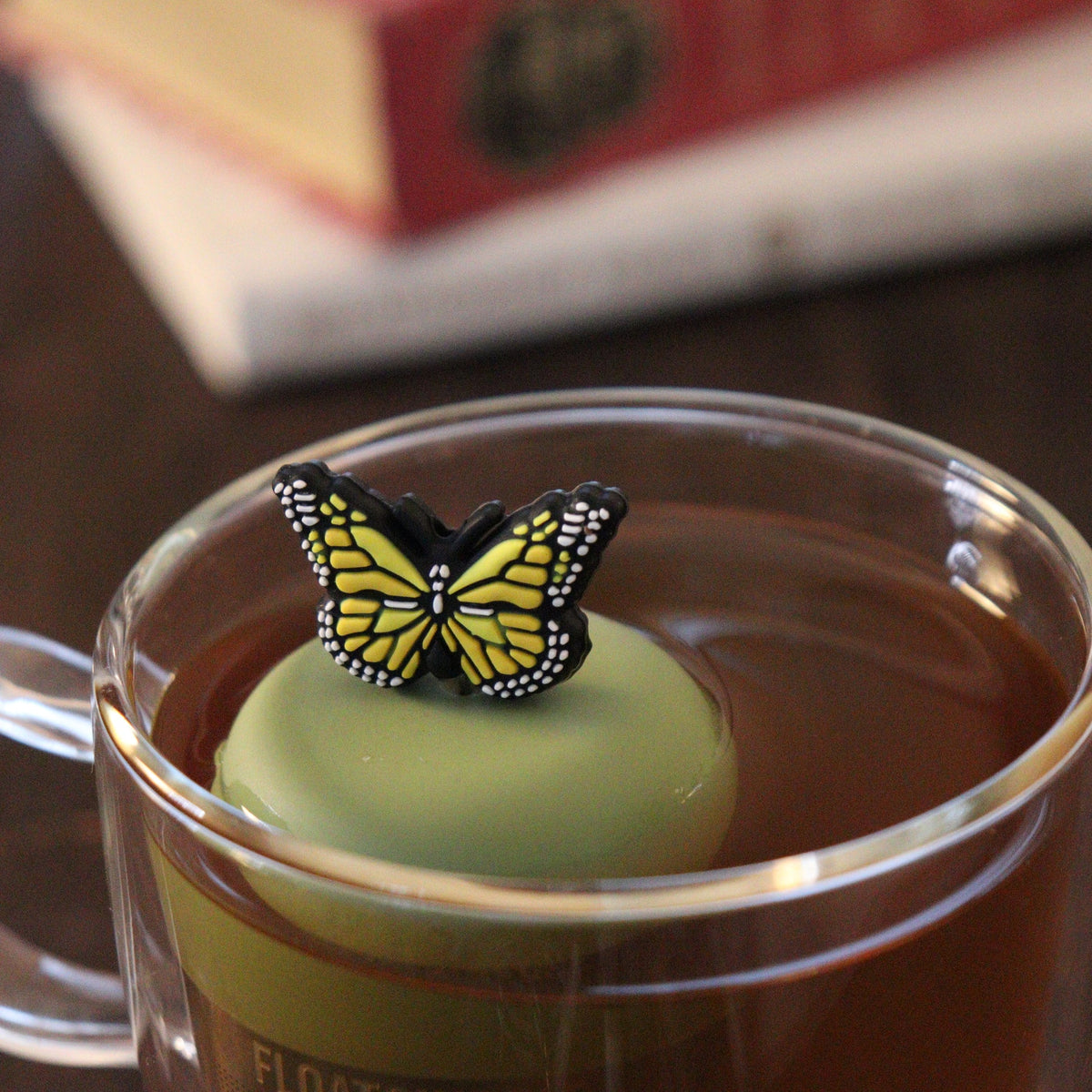 A clear glass mug of tea is paired with the Float2infuse Tea Infuser, featuring their Yellow Butterfly - Floating Tea Infuser, elegantly topped with a charming yellow and black butterfly decoration. This whimsical accessory sits gracefully against a backdrop of books.