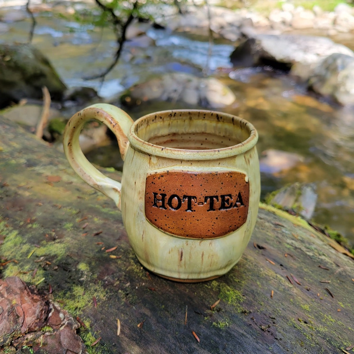 Handmade "HOT-TEA" pottery mug sitting outside on a rock by the river
