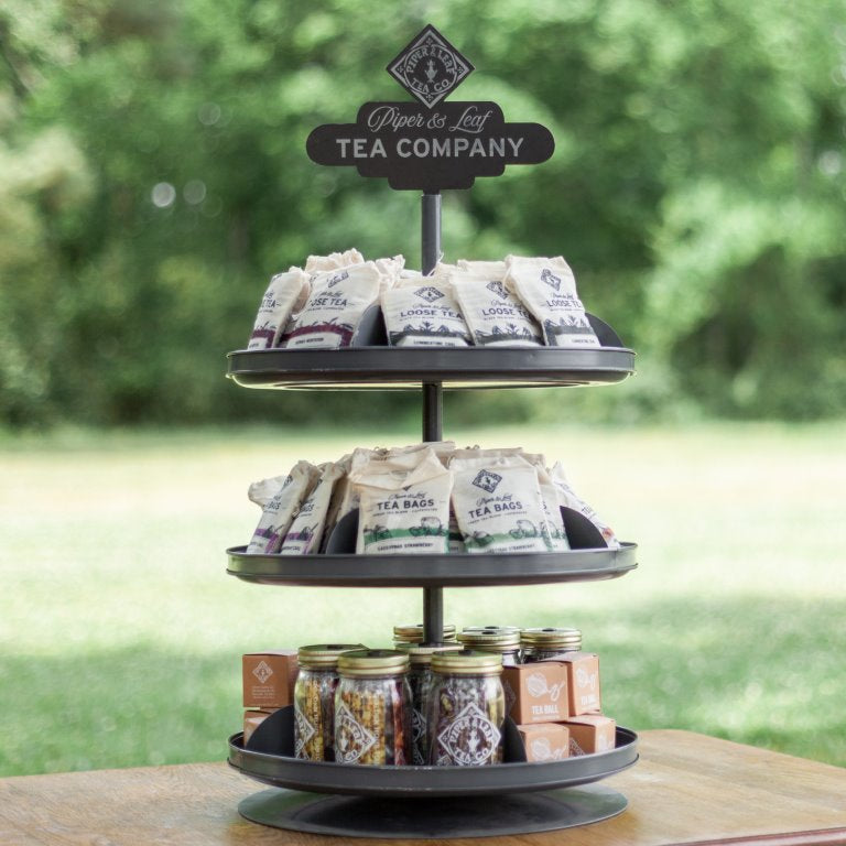 The 3-Tier Display Bundle - Classic Blends by Piper and Leaf Tea Co. showcases tea packets and small jars outdoors against a blurred green backdrop.