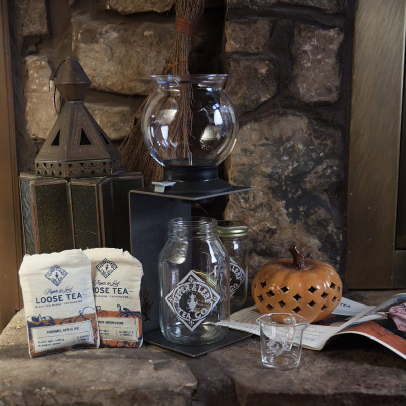 A deluxe brew kit with bags of Caramel Apple Pie and Pumpkin Moonshine loose leaf. The kit is surrounded by autumn decor: a pumpkin candle holder, an antique lantern, and a pine-needle broom.
