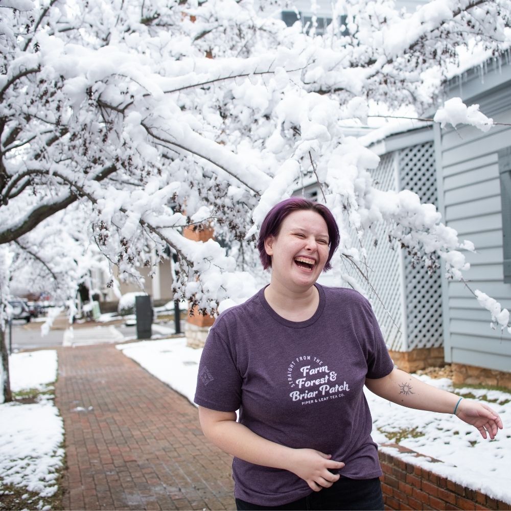 A woman laughing outside in the soft snow while wearing a "Farm, Forest, Briar Patch" Tea-Shirt by Piper & Leaf Tea Co.