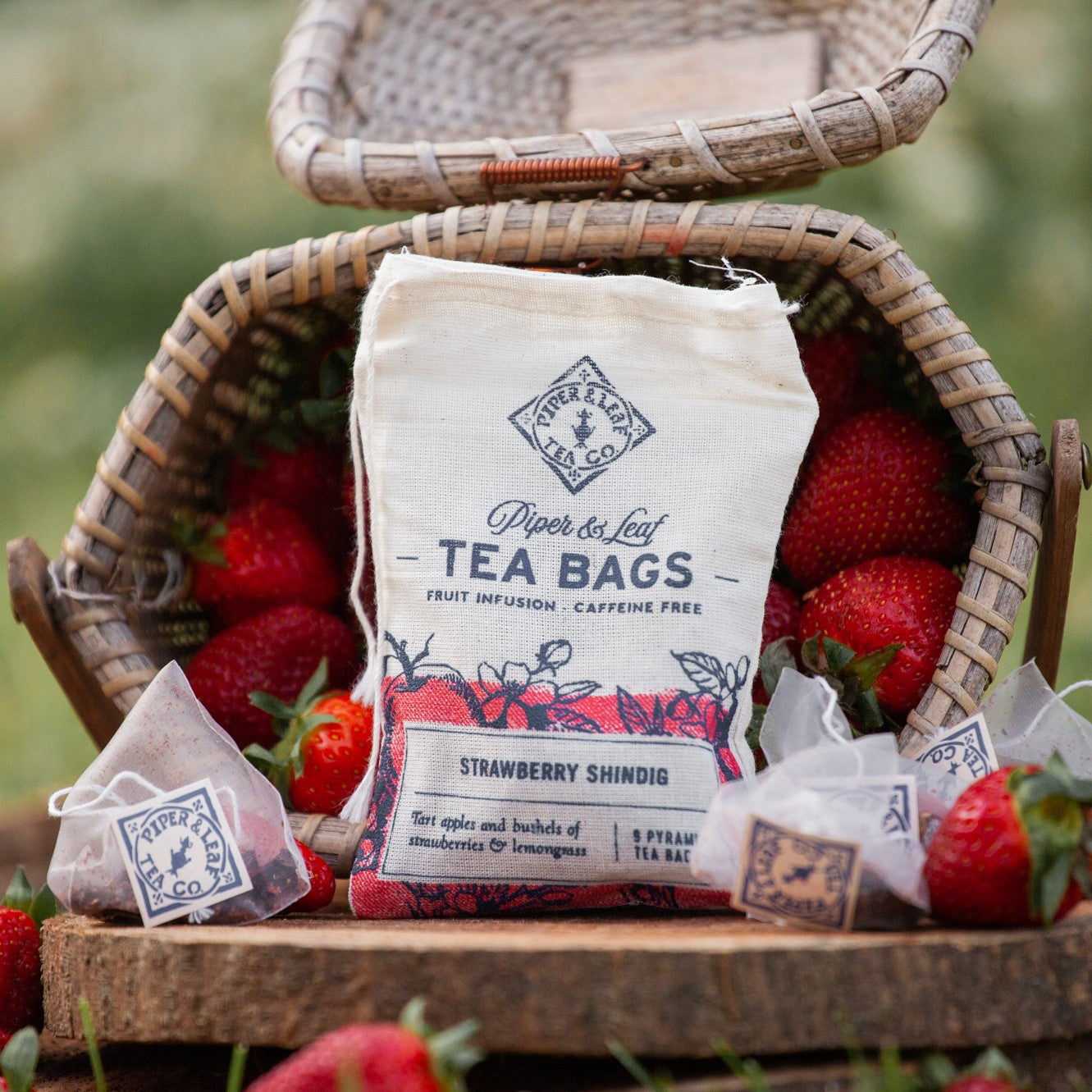 A muslin bag of 9ct sachets: Strawberry Shindig. The bag is resting amid tea bags in front of a basket overflowing with strawberries.