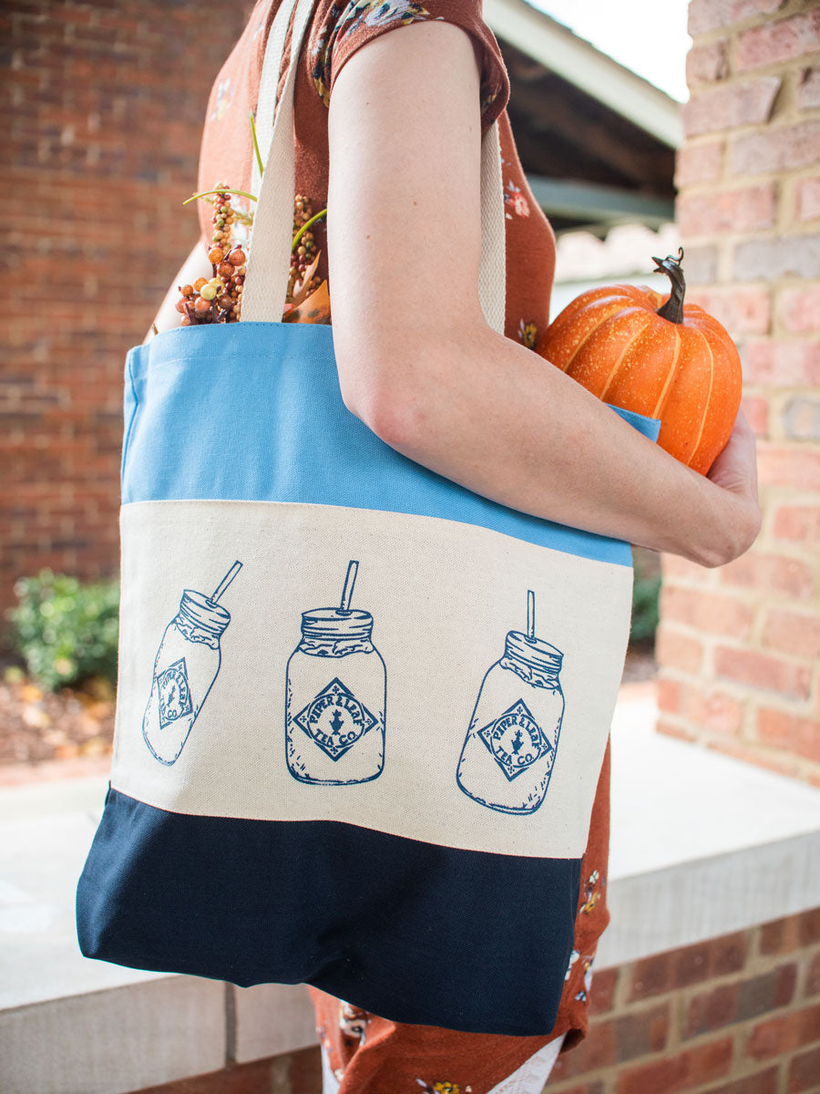 A woman wearing a blue and muslin tote bag over her shoulder. The bag is printed with mason jars and she is holding a pumpkin