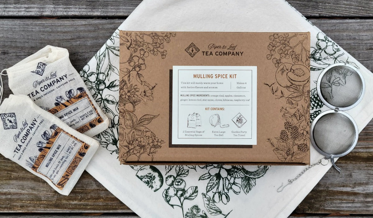 A Piper & Leaf Tea Co. branded Mulling Spices Brewing Kit box with intricate patterns, two loose leaf tea packets, and two tea balls, set on a wooden surface and a white cloth with botanical prints. This elegant kit makes for a perfect housewarming gift.