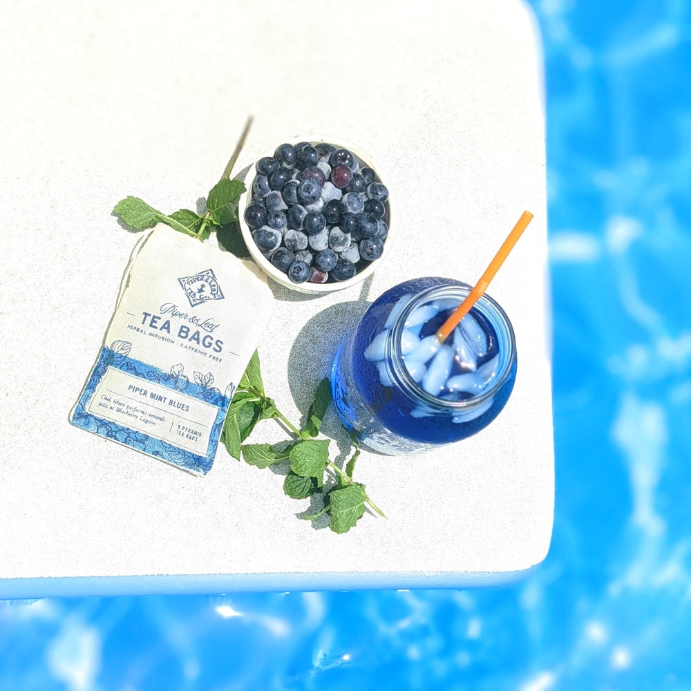 A top down view of an iced jar of Piper Mint Blues, its tea bags, a bowl of fresh blueberries, and a sprig of mint.