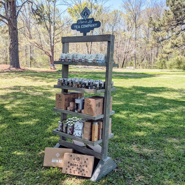 The Ladder Display Bundle - Classics by Piper and Leaf Tea Co. features sturdy shelves showcasing various tea products, including boxed brews and boba kits, set in a grassy area with trees in the background and enhanced by eye-catching promotional signage.