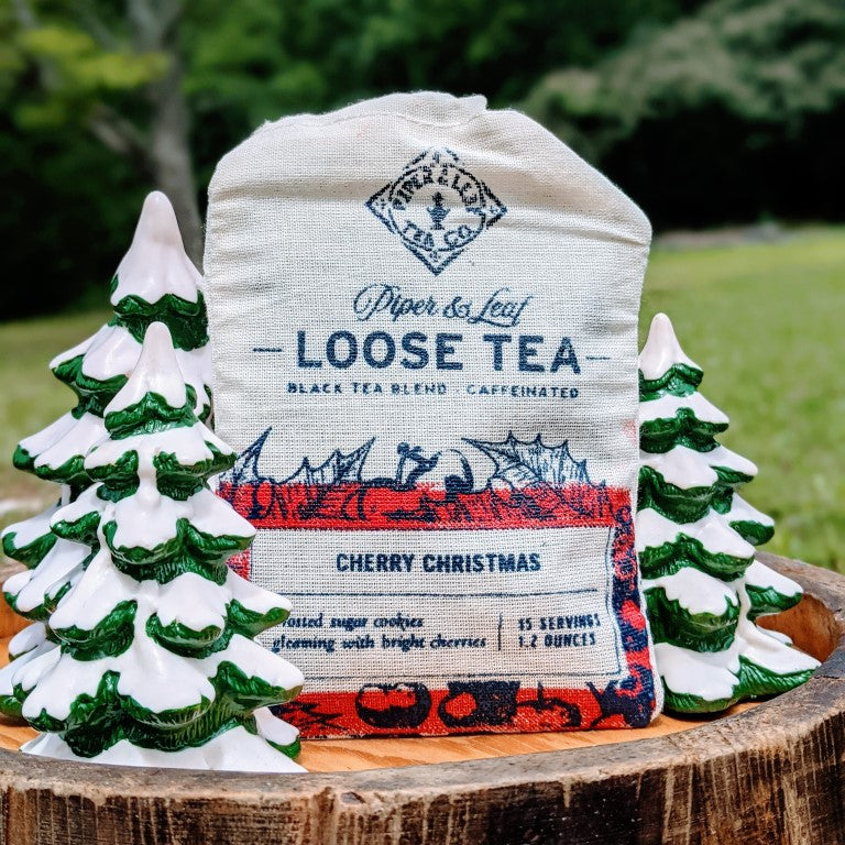 A "Piper & Leaf Tea Co." Cherry Christmas Muslin Bag of Loose Leaf Tea - 15 Servings is displayed outdoors surrounded by small decorative ceramic trees. The label notes it is a Ceylon Black tea blend with 15 servings per bag.