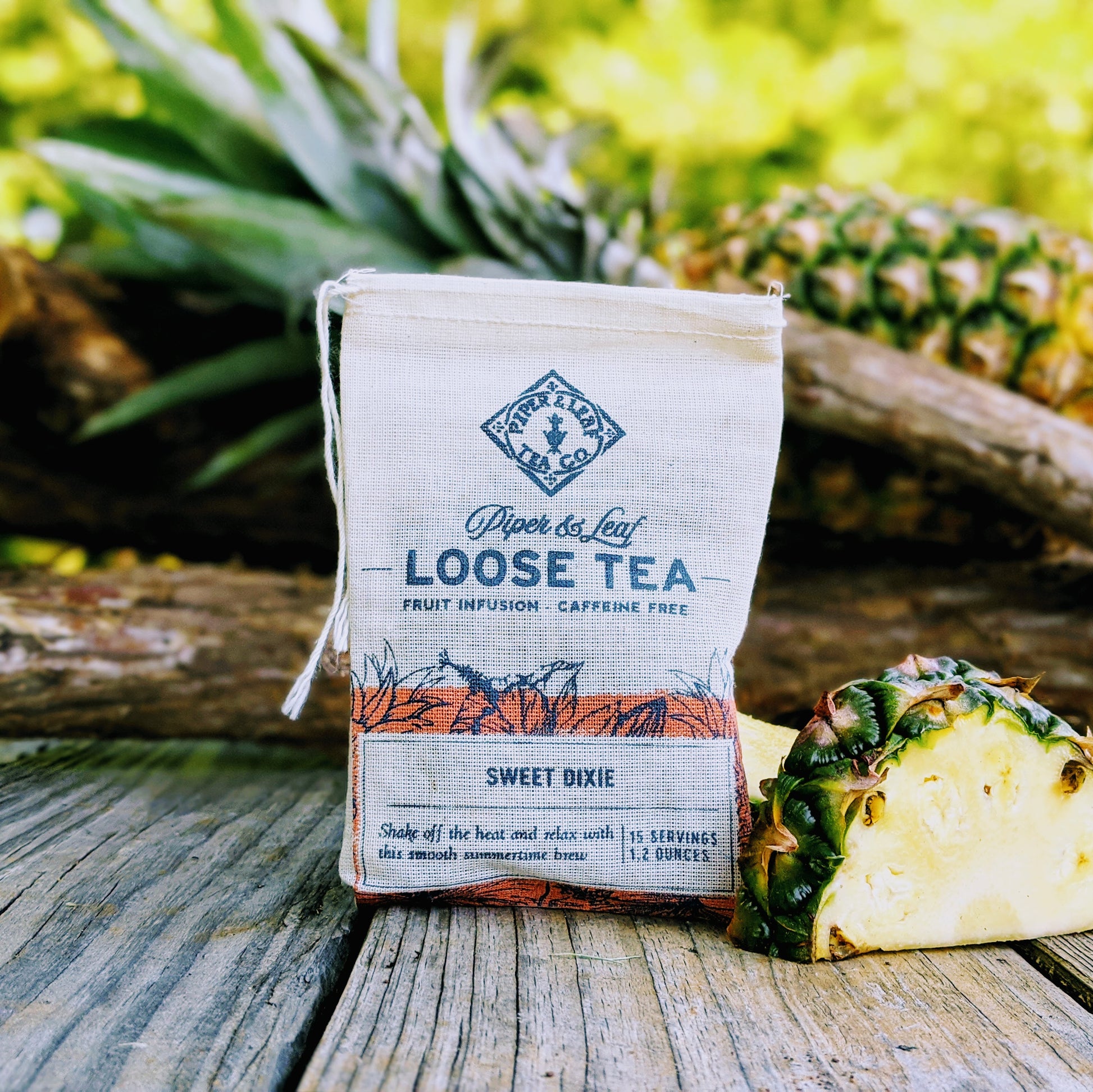 A packet of Sweet Dixie Muslin Bag of loose leaf fruit blend tea from Piper & Leaf Tea Co., labeled caffeine-free, displayed next to a fresh pineapple slice on a wooden surface.