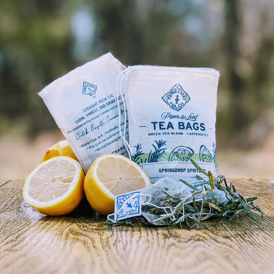 Piper & Leaf Tea Co.'s Springdrop Spritzer 9ct Tea Bags in Muslin with lemons and rosemary on a wooden table, offering a refreshing taste.