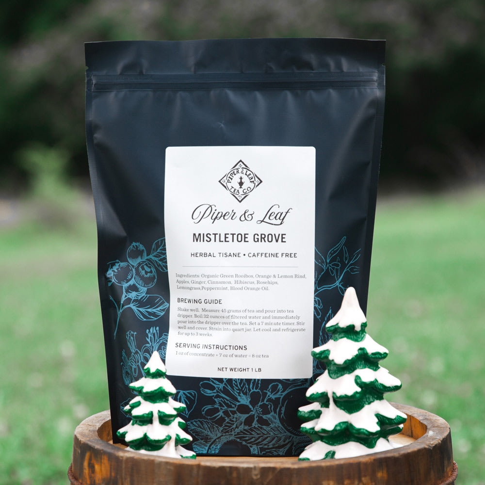 A black packet labeled "Piper & Leaf Tea Co. Mistletoe Grove Pound Bag - 190 servings" is displayed outdoors, with two small ceramic Christmas trees in the foreground. This caffeine-free, holiday favorite loose leaf tea adds a festive touch to any setting.