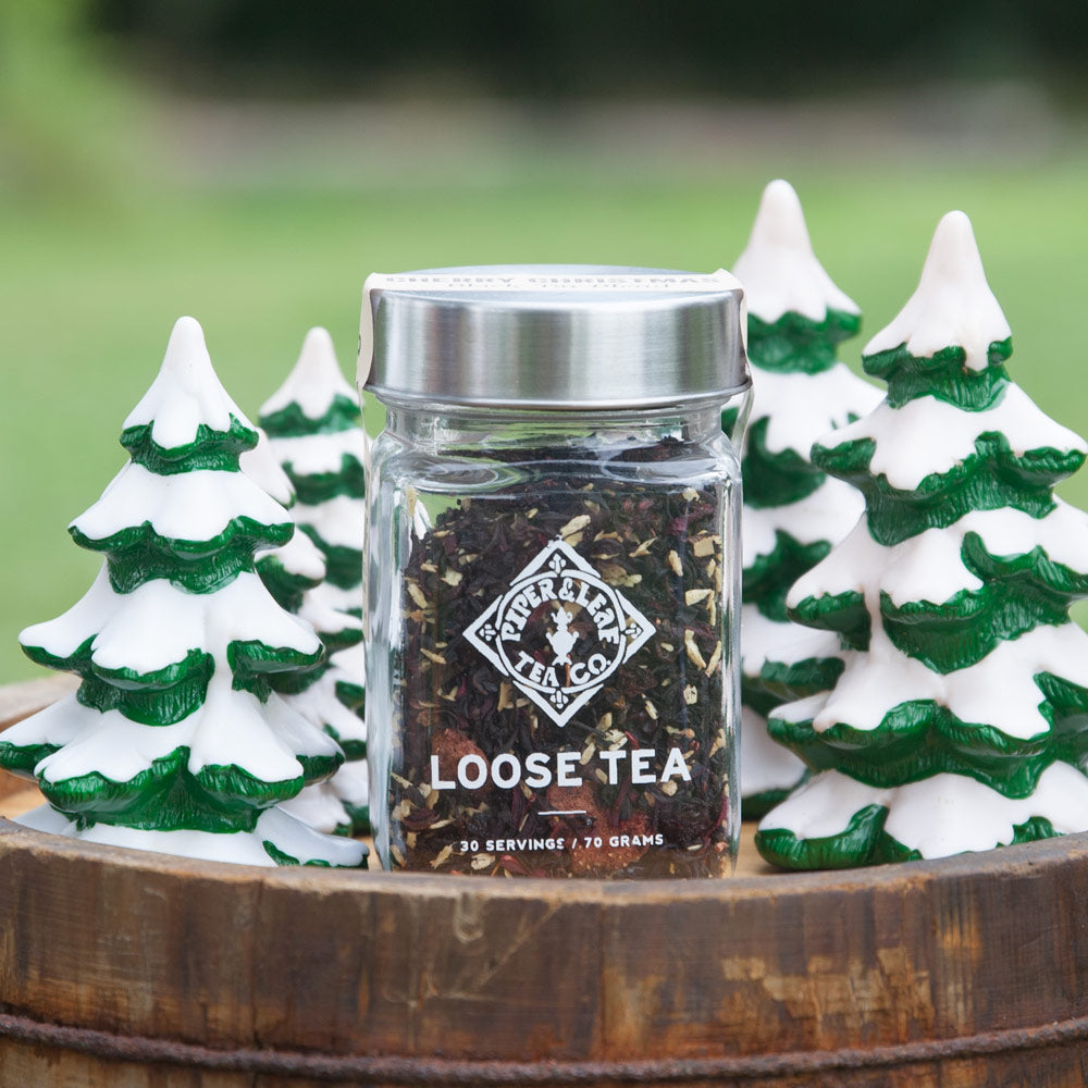 A jar of Cherry Christmas Glass Jar of Loose Leaf Tea - 30 Servings from Piper & Leaf Tea Co. is placed on a wooden surface, surrounded by miniature green and white tree decorations.