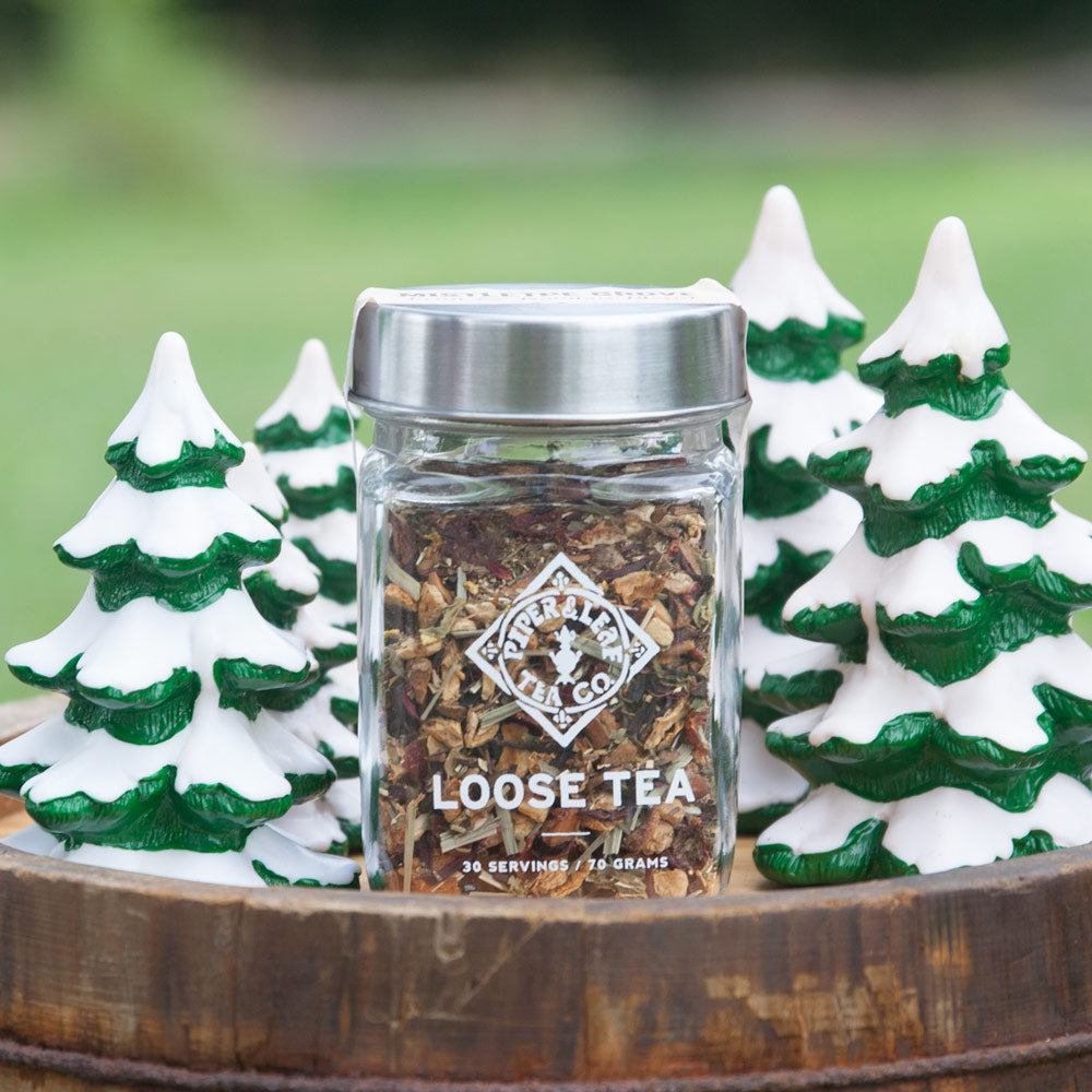 A jar of Mistletoe Grove Glass Jar of Loose Leaf Tea - 30 Servings by Piper & Leaf Tea Co. is displayed on a wooden surface, surrounded by small decorative trees with snow-topped green foliage, promising a caffeine-free delight for the season.