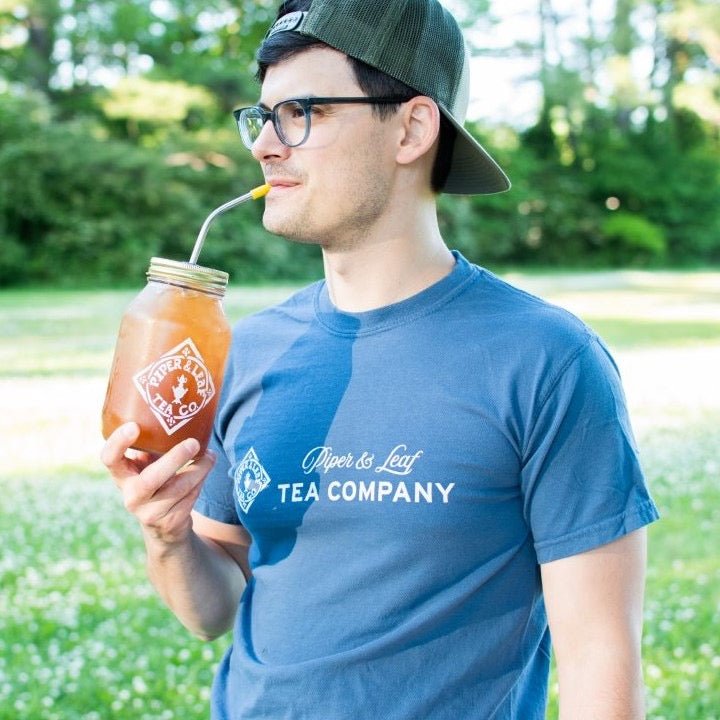 A man in glasses sips from a quart jar of tea while wearing a blue Piper & Leaf Tea Company tee-shirt