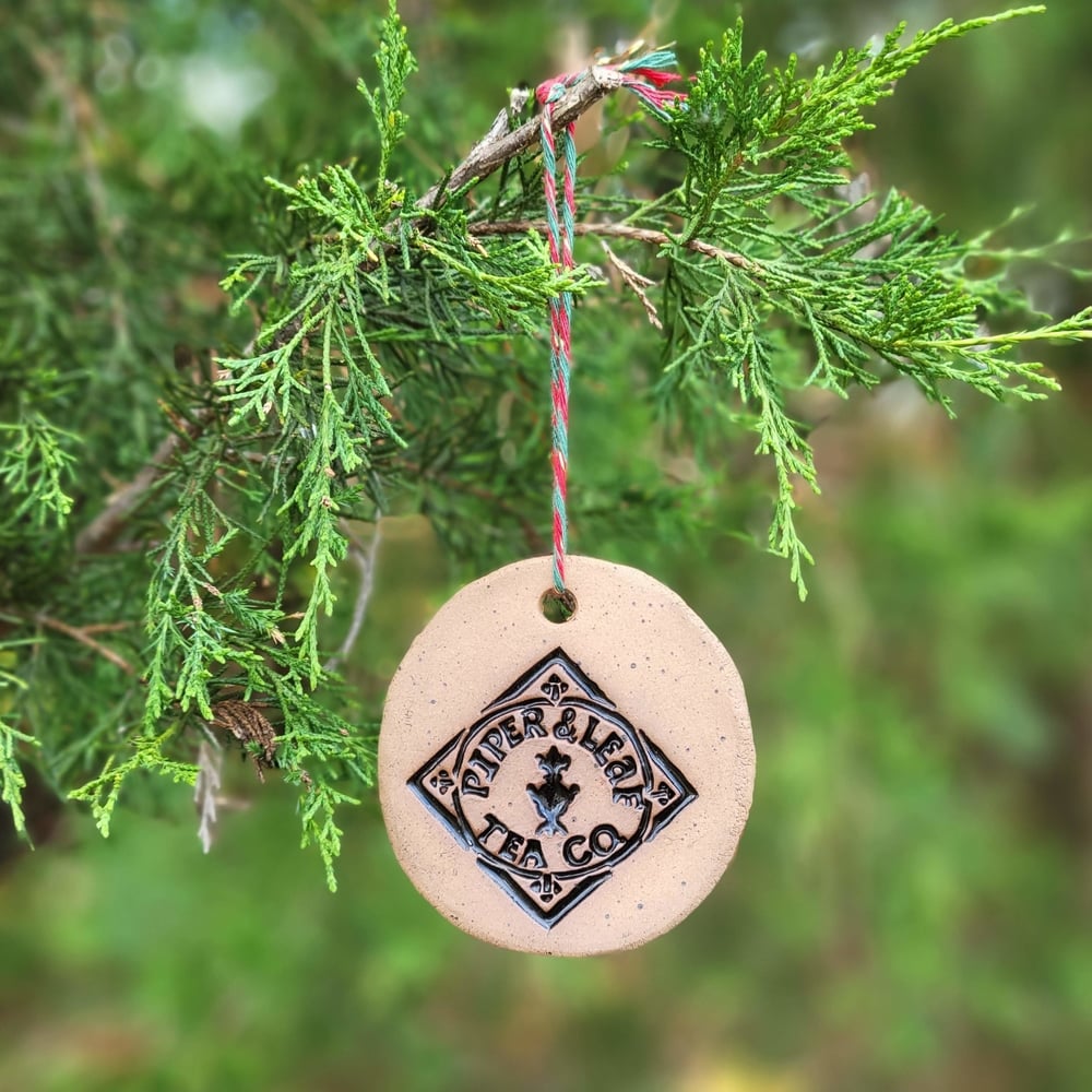 A round pottery ornament hanging from a fir tree - the tan ornament is engraved with a black Piper & Leaf diamond logo and hung by red and green string.