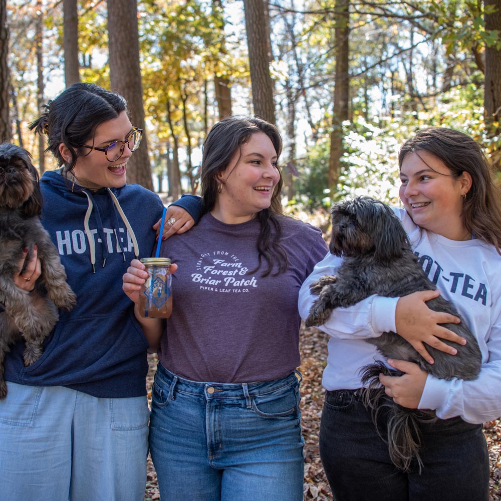 Three girls standing around with dogs and jars of tea. The girl on the left is in a blue "HOT-TEA" sweatshirt, the girl in the middle wears a purple "farm, forest, & briar patch" shirt, and the girl on the right has a "Hot-tea" white shirt on