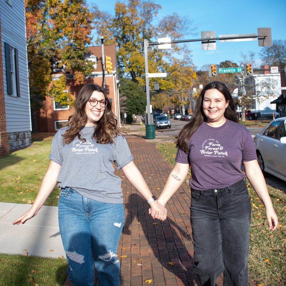 Two women holding hands on a sidewalk, wearing Piper & Leaf Tea Co.'s "Farm, Forest, Briar Patch" Tea-Shirt.