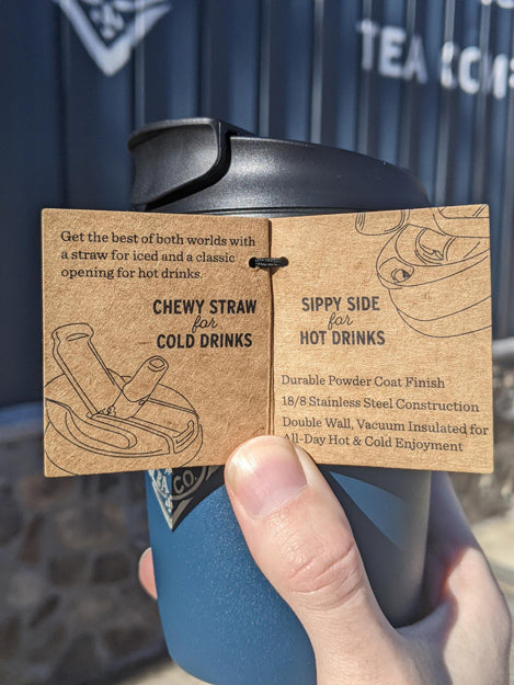 Right hand holding a blue Piper & Leaf travel cup with tag open showing pictures of the chewy straw and sippy side capabilities for iced and hot drinks.