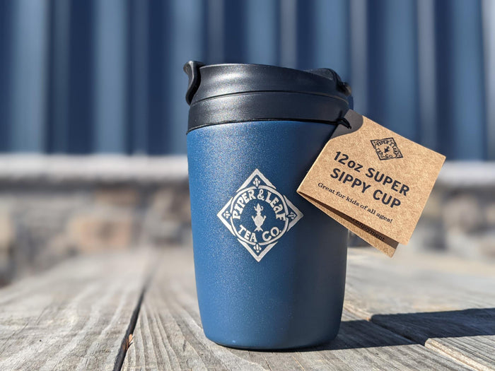 A blue Piper & Leaf logoed travel cup with paper tag sitting on wooden table outside in sunshine