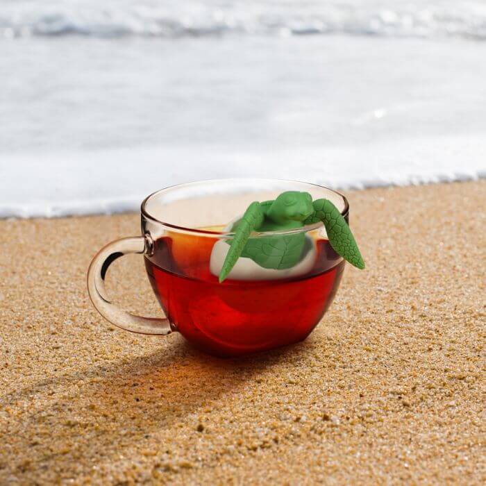 A sea turtle shaped Fred-brand tea strainer in a glass tea cup on the beach: the Tea Turtle
